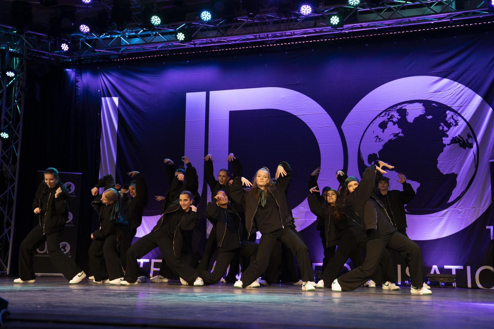 Tanzgruppe in schwarzen Outfits f&uuml;hrt eine choreografierte Darbietung auf einer B&uuml;hne mit JDO-Logo vor.