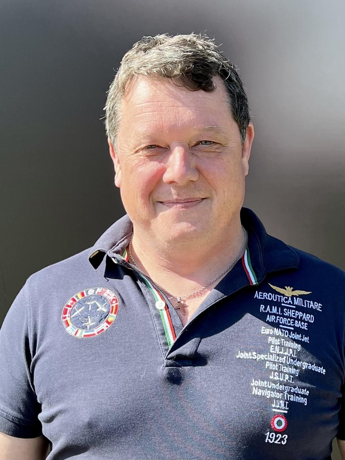 L&auml;chelnder Mann mit kurzen Haaren, tr&auml;gt ein dunkelblaues Poloshirt mit Abzeichen und steht vor einem neutralen Hintergrund.