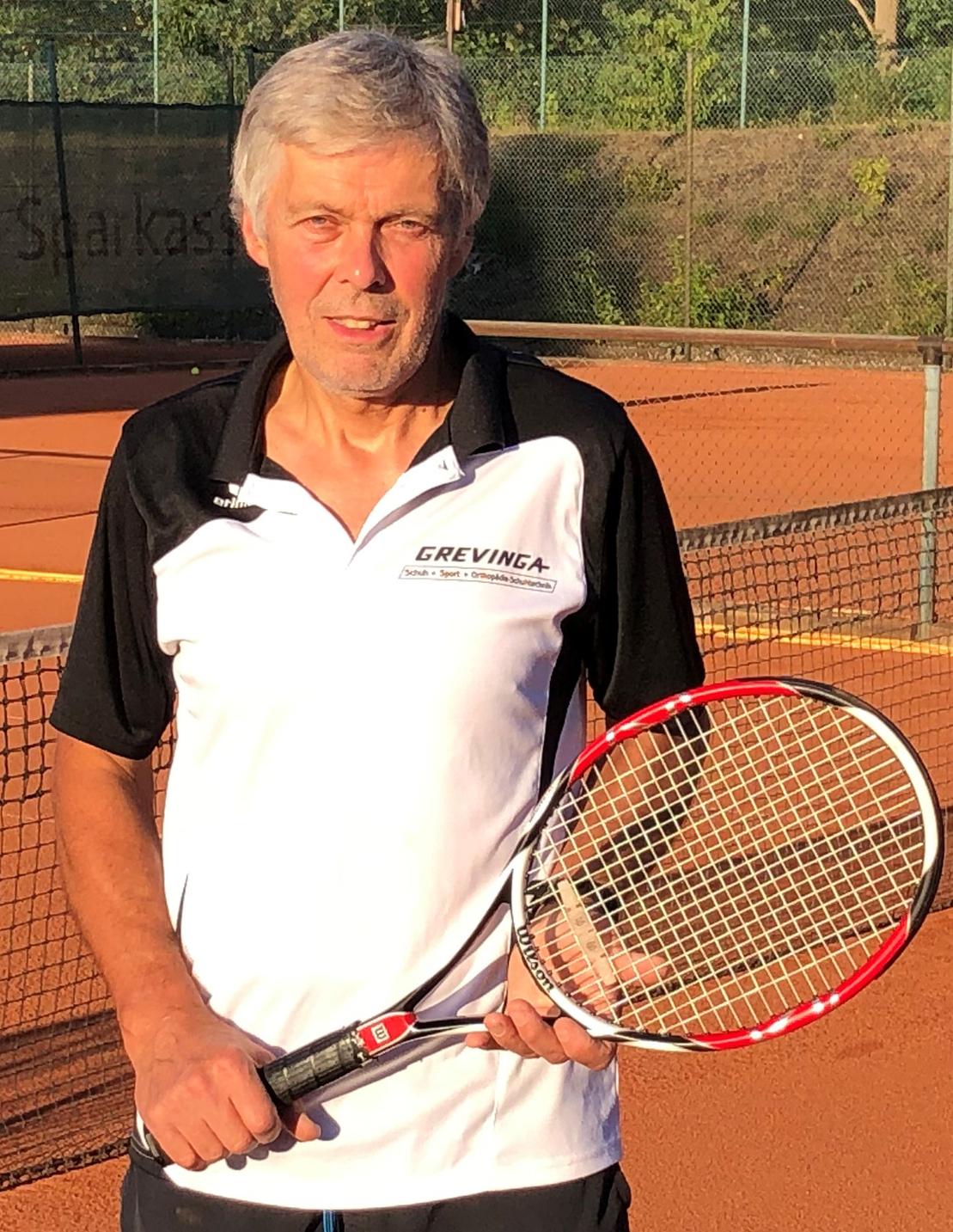 Mann h&auml;lt einen Tennisschl&auml;ger auf einem Sandplatz, im Hintergrund sind Tennisnetze und B&auml;ume sichtbar.