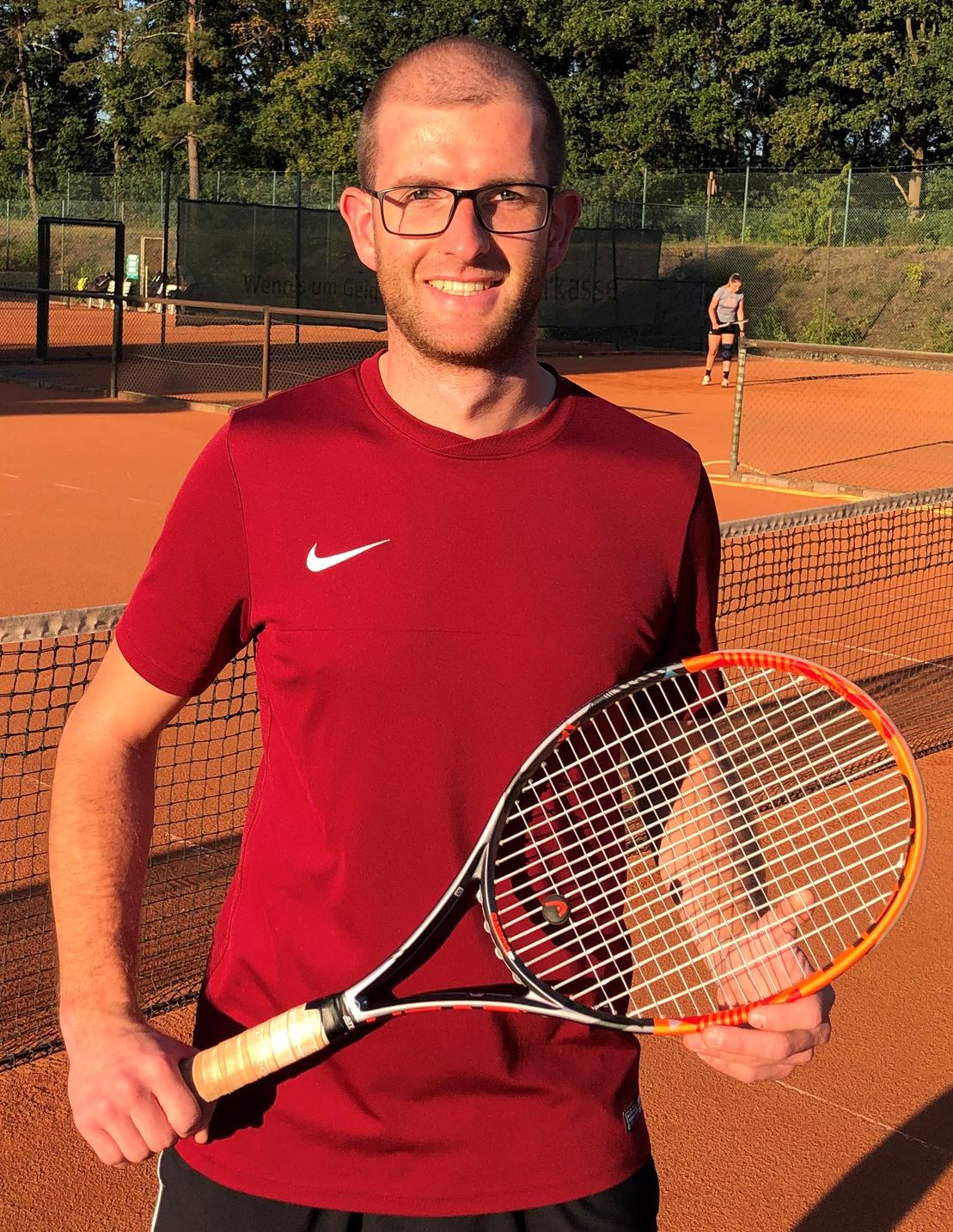 Mann in rotem Shirt mit Brille h&auml;lt einen Tennis-Schl&auml;ger auf einem Tennisplatz im Hintergrund.