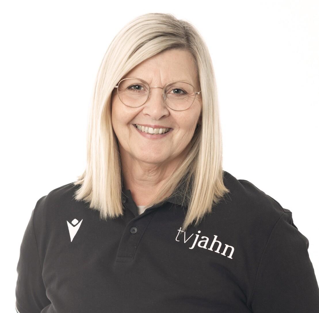 Portrait einer lächelnden Frau mit blonden, schulterlangen Haaren, Trägershirt mit Logo „tvjahn“ und Brille.