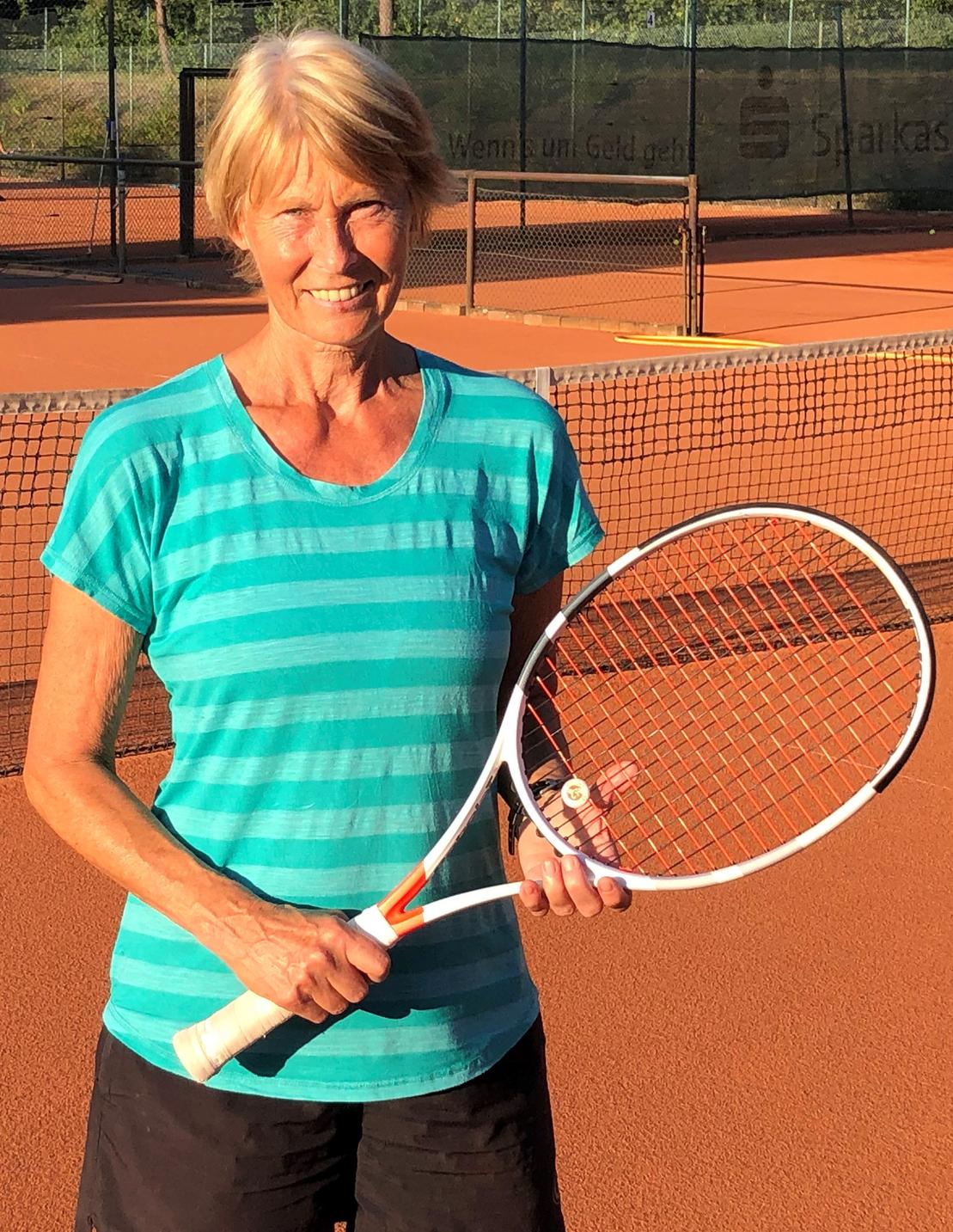 L&auml;chelnde Frau h&auml;lt einen Tennisschl&auml;ger auf einem Tennisplatz mit orangem Belag und Netz im Hintergrund.