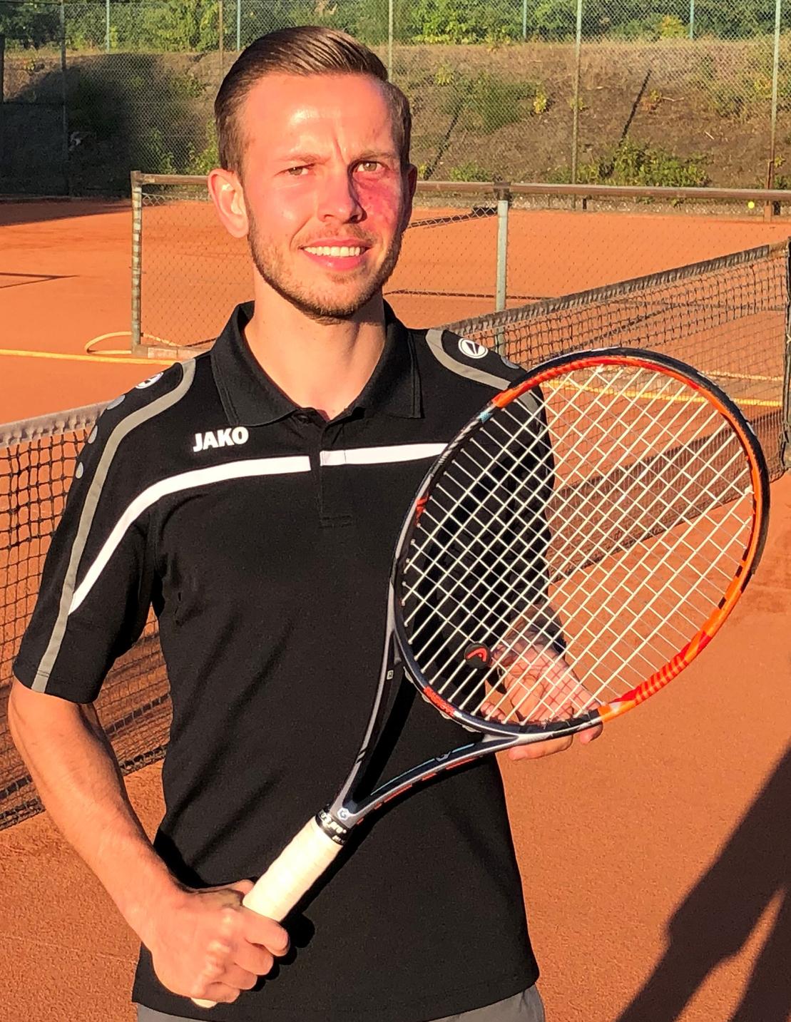 Ein junger Mann in einem schwarzen Poloshirt h&auml;lt einen Tennisschl&auml;ger auf einem Tennisplatz mit Sandbelag.