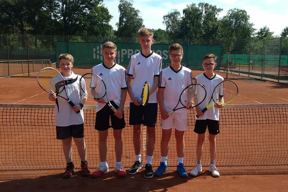 F&uuml;nf Jugendliche mit Tennisschl&auml;gern stehen l&auml;chelnd an einem Tennisplatz hinter dem Netz.