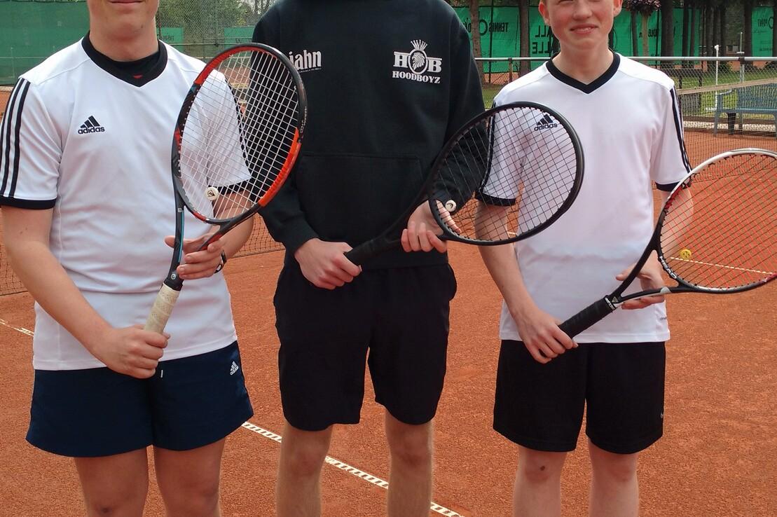Drei Jugendliche stehen auf einem Tennisplatz, jeder h&auml;lt einen Tennisschl&auml;ger in der Hand, im Hintergrund B&auml;ume.
