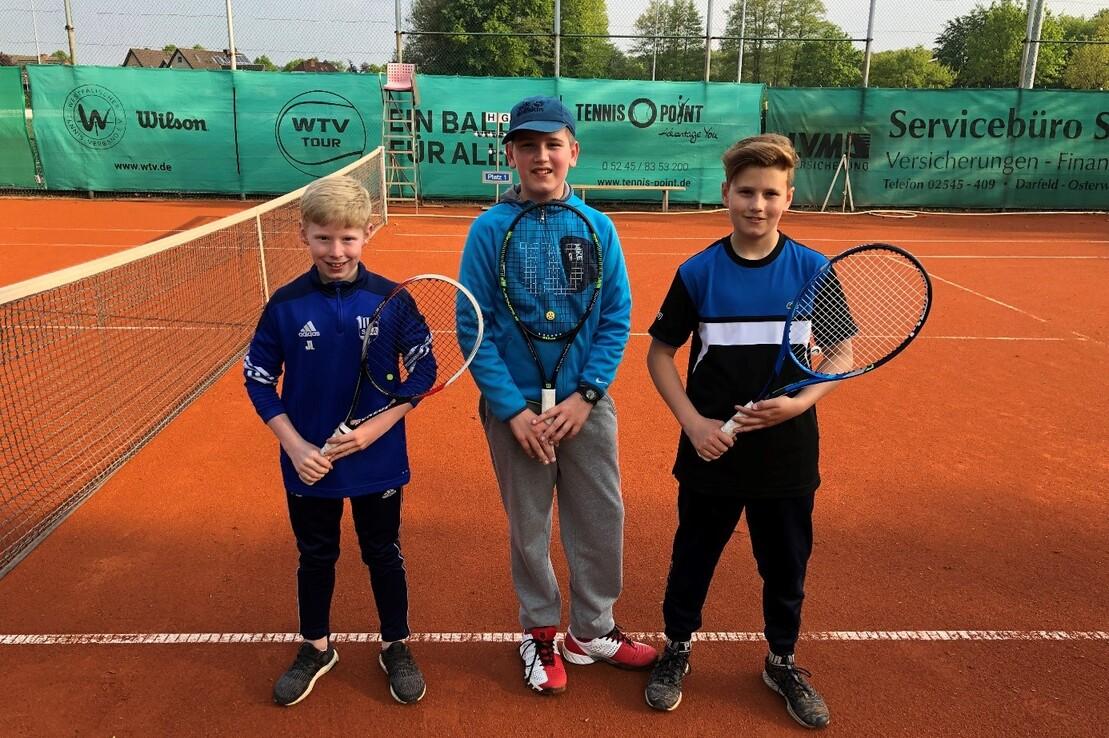 Drei Kinder stehen auf einem Tennisplatz, halten Tennisschl&auml;ger und posieren vor einem Netz.