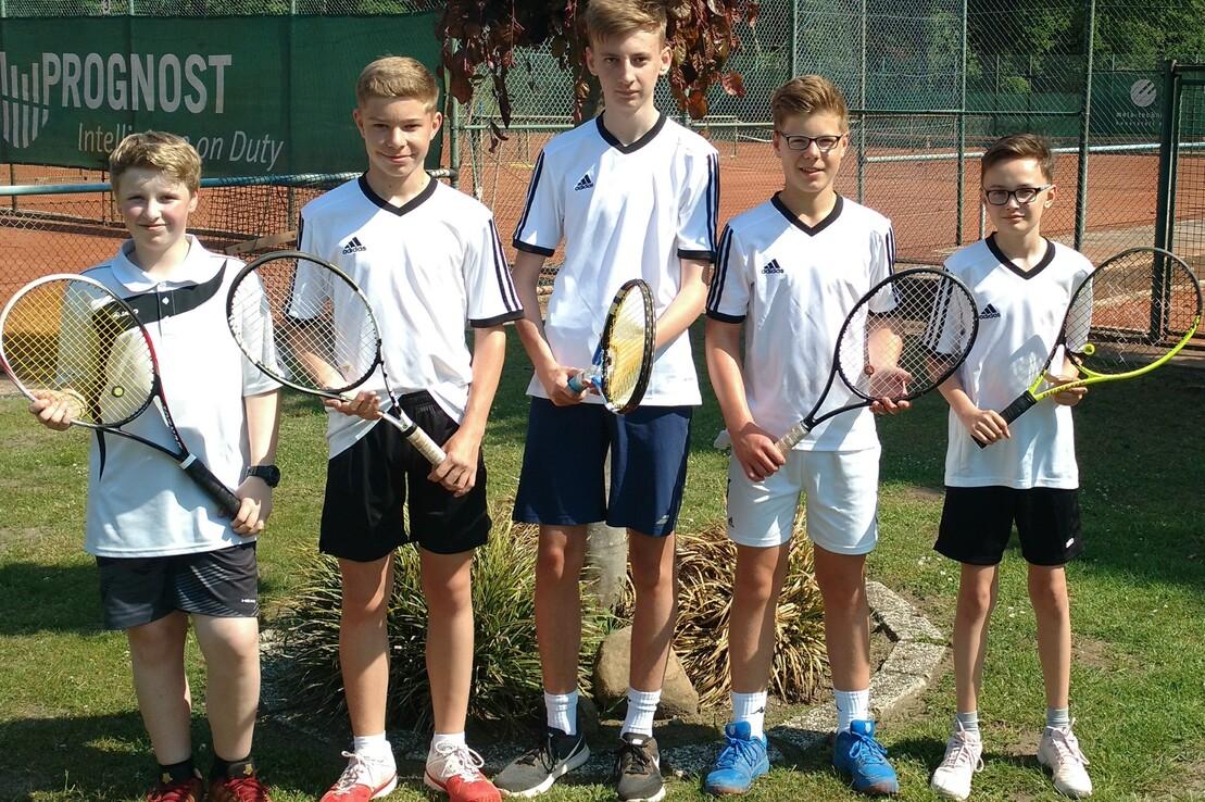F&uuml;nf Jugendliche mit Tennisschl&auml;gern stehen l&auml;chelnd auf einem Tennisplatz vor einer gr&uuml;nen Werbetafel.