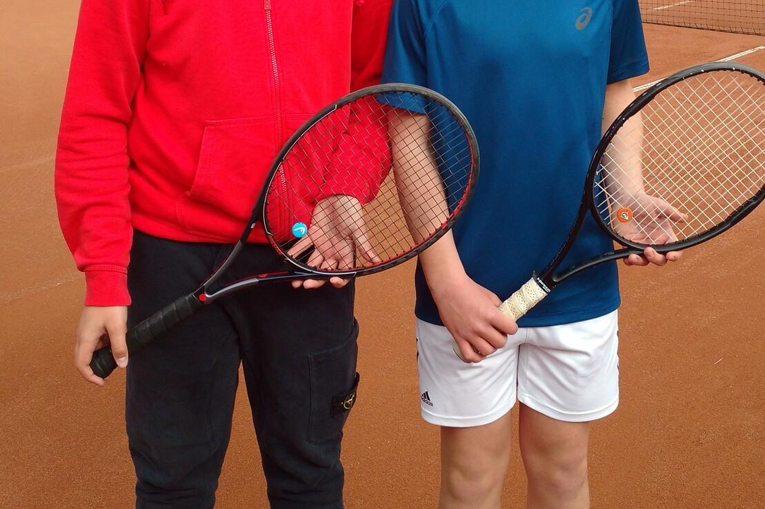 Zwei Jungen mit Tennisschl&auml;gern stehen auf einem Tennisplatz und l&auml;cheln in die Kamera.