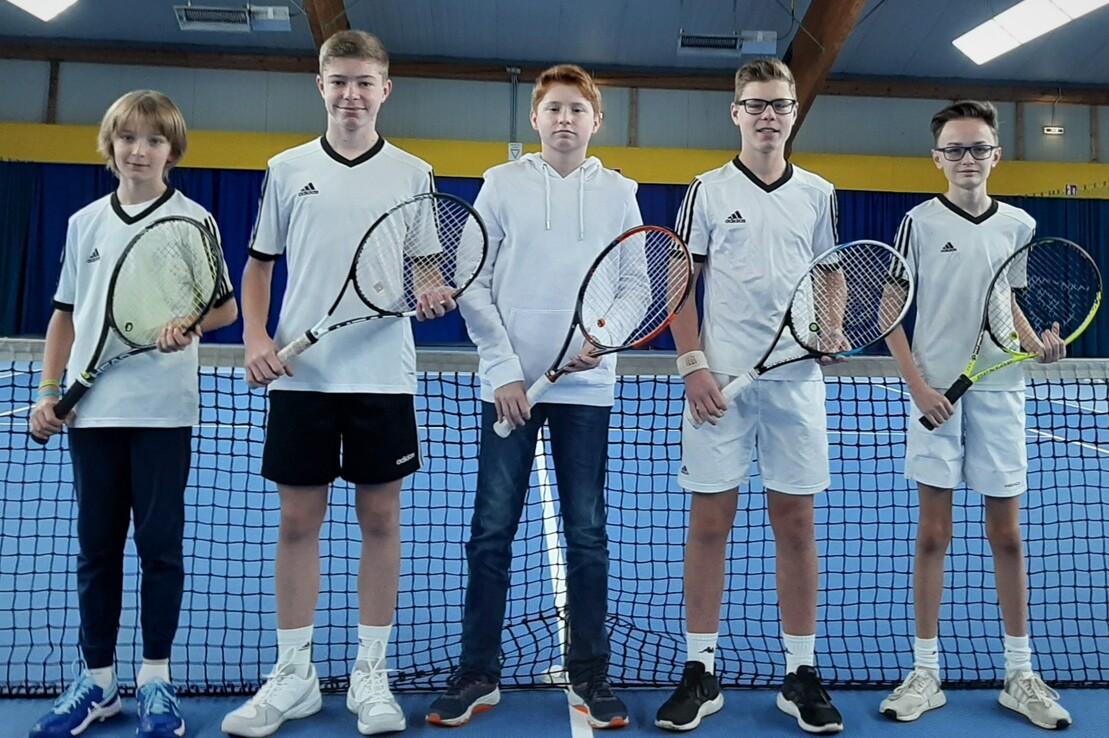 F&uuml;nf Jugendliche in wei&szlig;er Sportkleidung stehen vor einem Tennisnetz in einer Halle, jeder mit einem Tennisschl&auml;ger.