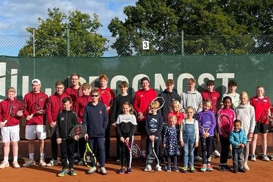 Gruppe von Jugendlichen und Erwachsenen in roten Sweatshirts mit Tennisschlägern auf einem Tennisplatz vor einer Wand.