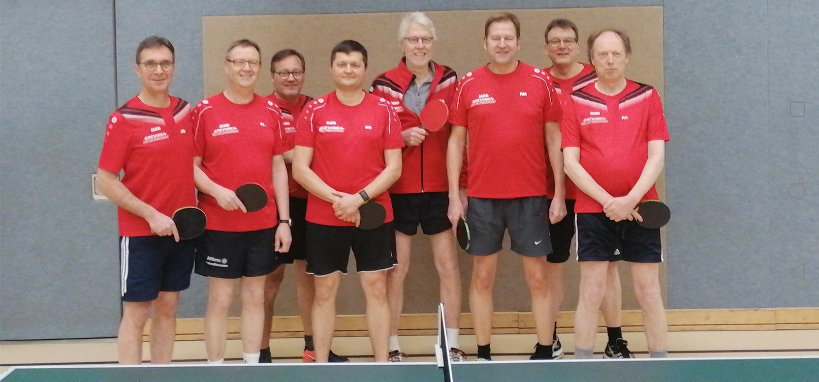 Gruppenfoto von acht Tischtennisspielern in roten Trikots vor einem Tischtennistisch in einer Sporthalle.