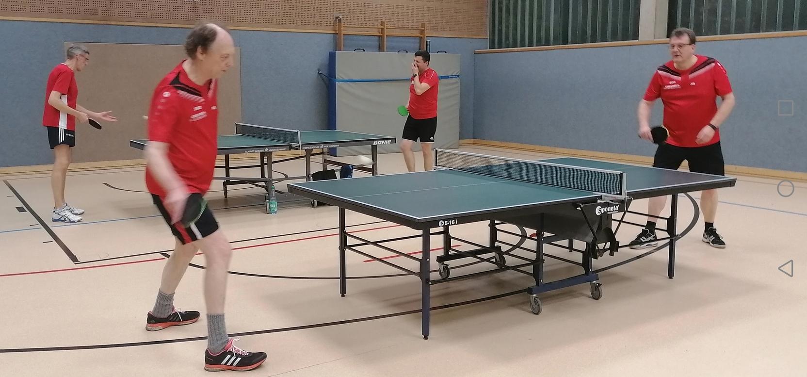 Vier Tischtennisspieler in roten Shirts trainieren an zwei Tischtennisplatten in einer Sporthalle.