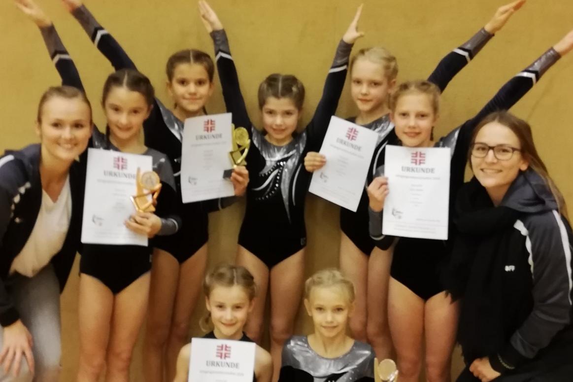 Gruppe von sieben Turnerinnen und Trainerinnen, die stolz mit Medaillen und Urkunden posieren. Sporthalle im Hintergrund.