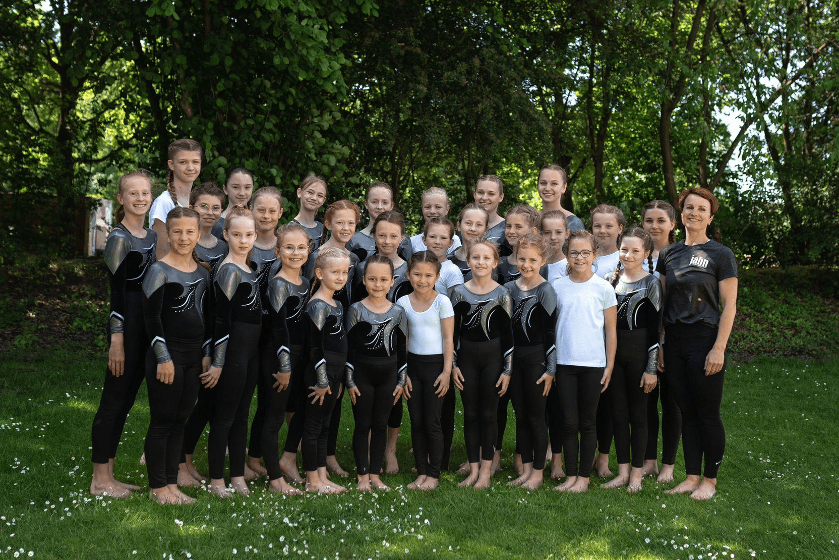 Gruppe von Turnerinnen in schwarz-silbernen Anz&uuml;gen sowie einer Trainerin in T-Shirt, im Freien auf einer Wiese.