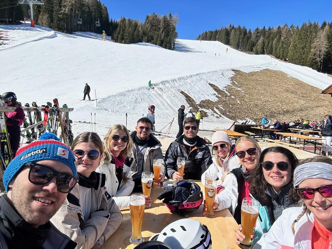 Gruppe von acht Personen sitzt an einem Tisch mit Getr&auml;nken, Blick auf eine schneebedeckte Piste und Wald im Hintergrund.