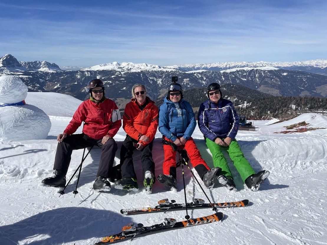 Vier Skifahrer sitzen auf Schneelage mit Bergen im Hintergrund, Skiausr&uuml;stungen liegen vor ihnen.