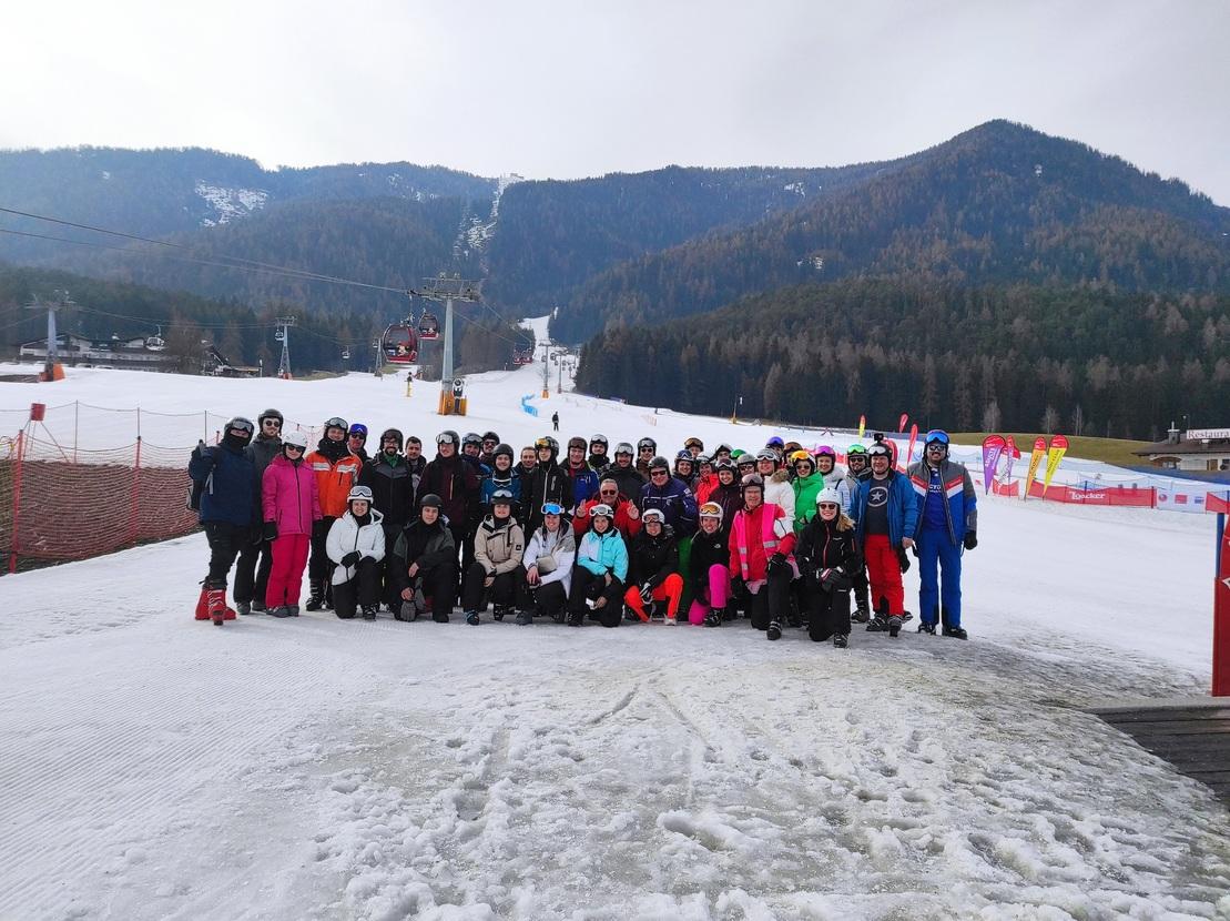 Gruppenfoto von etwa 60 Skifahrern in Winterkleidung auf einer verschneiten Piste vor Bergen und Skiliften.