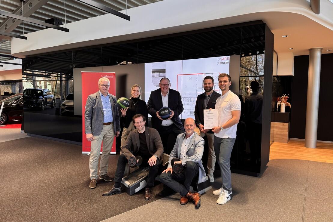 Gruppe von sechs Personen im Autohaus, posieren mit mehreren B&auml;llen und einer Urkunde vor einem digitalen Bildschirm.