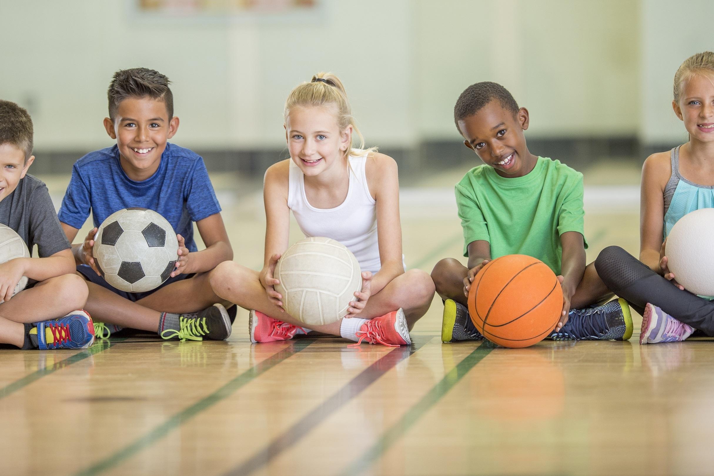 Gruppe von f&uuml;nf Kindern sitzt auf einem Sportboden, jeder h&auml;lt einen Ball: Fu&szlig;ball, Volleyball, Basketball und zwei Joker.