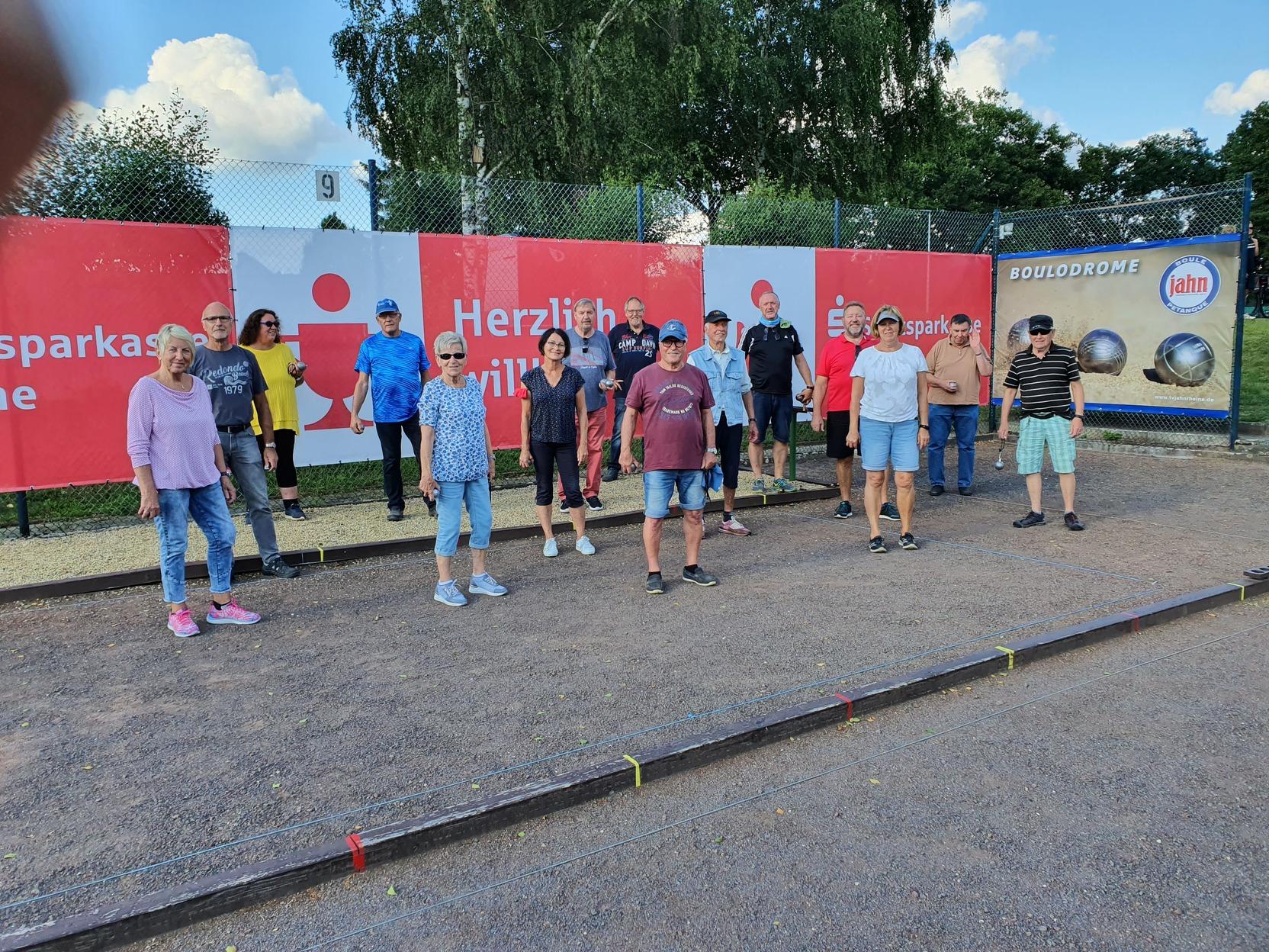 Gruppierung von Menschen auf einem Bouleplatz, mit Werbebannern im Hintergrund und einer klaren, sonnigen Himmelsszene.