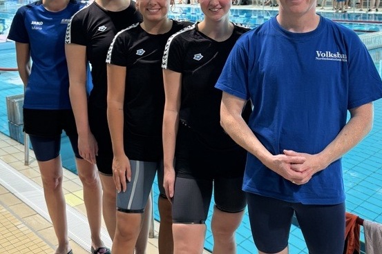 F&uuml;nf Schwimmer in Badebekleidung stehen am Beckenrand eines Schwimmbades, l&auml;chelnd, mit Wasser im Hintergrund.