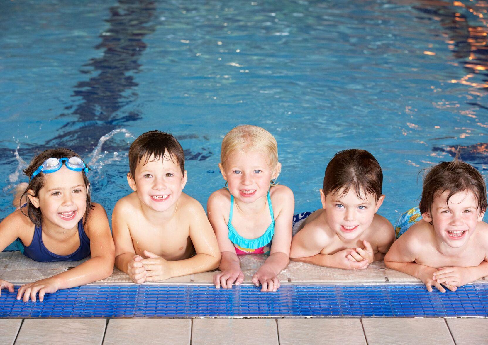 F&uuml;nf Kinder l&auml;cheln am Beckenrand eines Schwimmbeckens, ihre Arme auf dem Rand abgest&uuml;tzt.