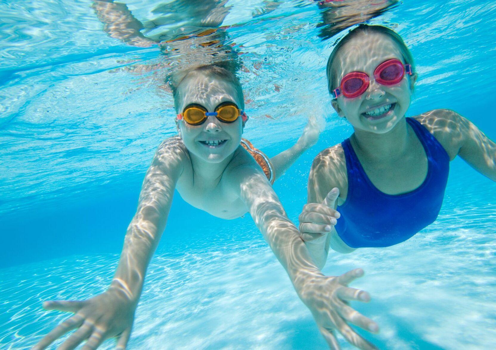 Zwei Kinder schwimmen unter Wasser, l&auml;cheln und tragen bunte Schwimmbrillen in einem blauen Schwimmbecken.