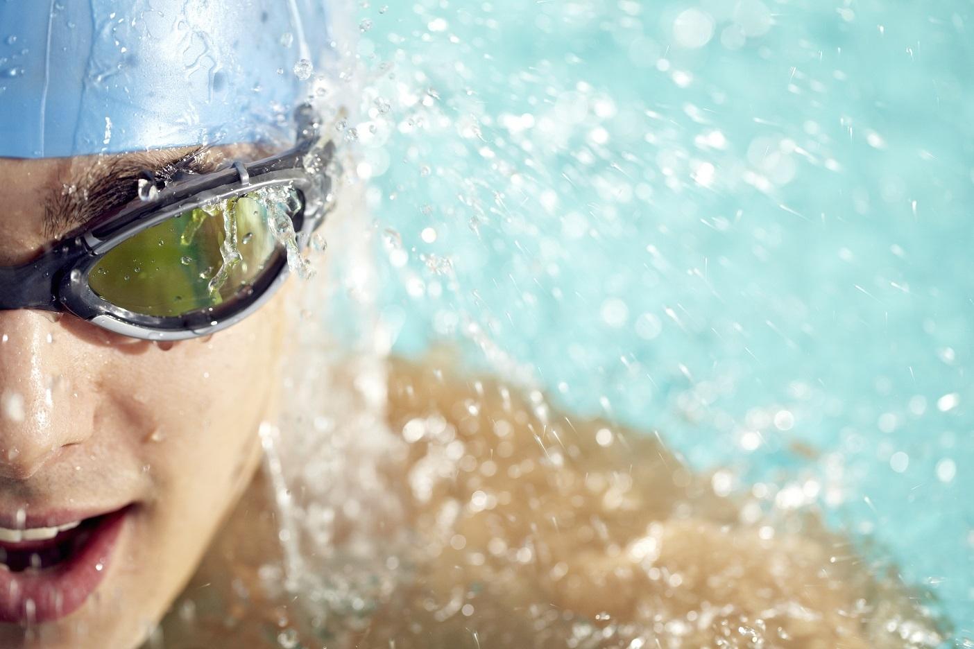 Nahaufnahme eines Schwimmers mit Badekappe und Schwimmbrille, Wasser spritzt um sein Gesicht.