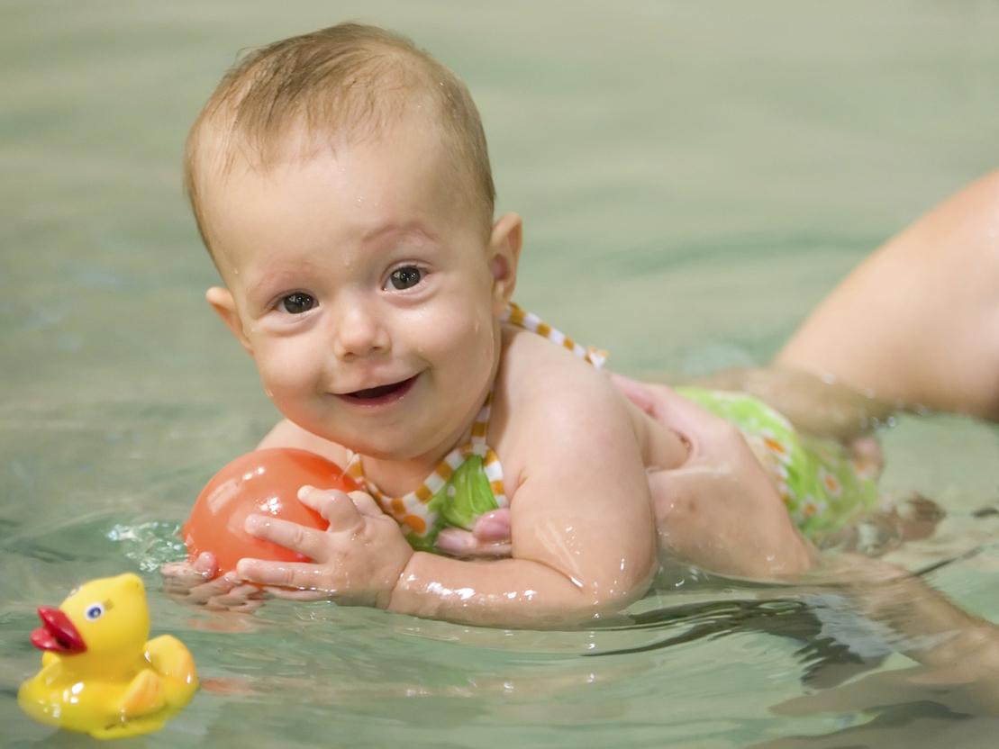 L&auml;chelndes Baby im Wasser mit einem orangefarbenen Ball, neben einem gelben Gummientchen schwimmend.