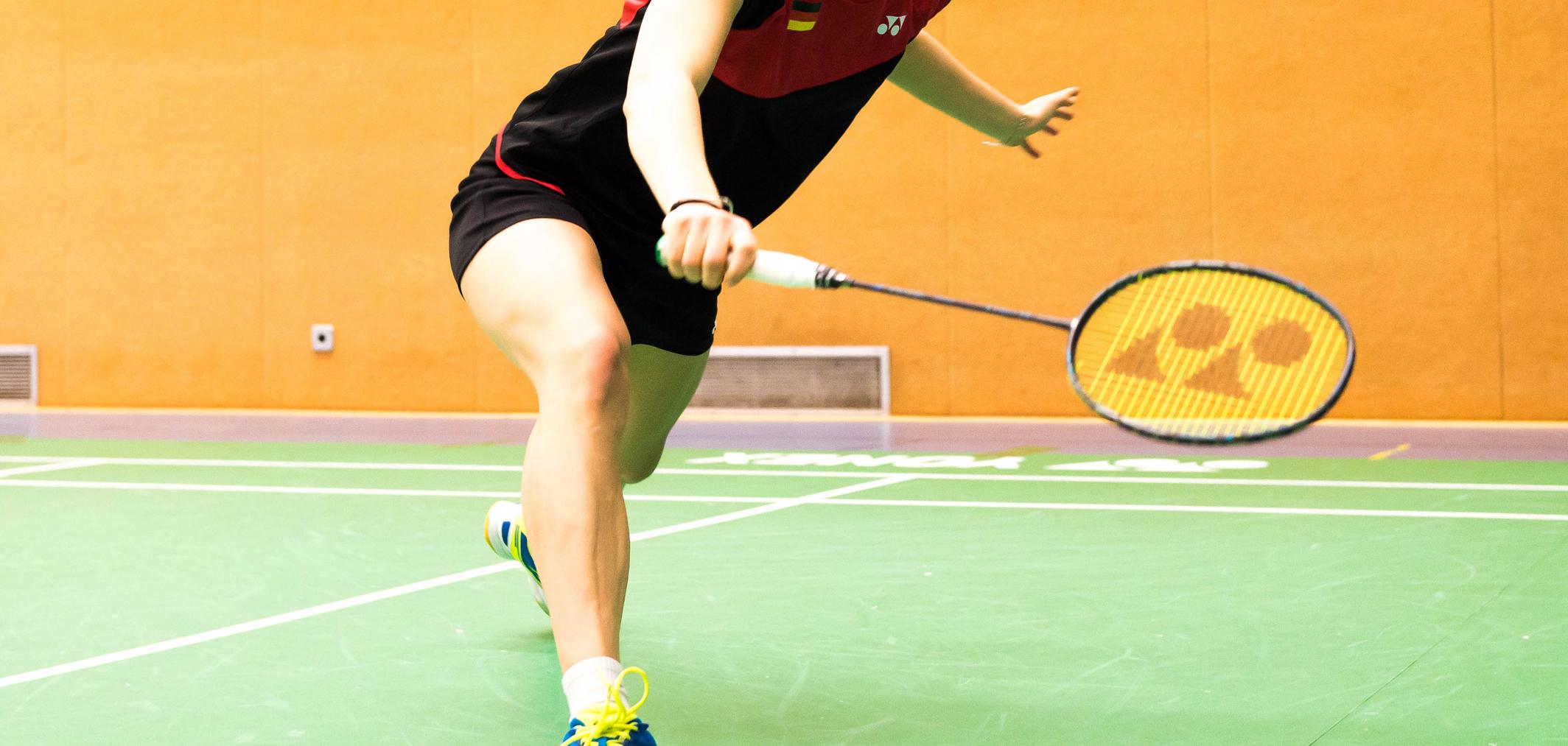 Spielerin schlägt einen Badmintonball auf einer grünen Spielfläche in einer Sporthalle mit orangefarbenen Wänden.