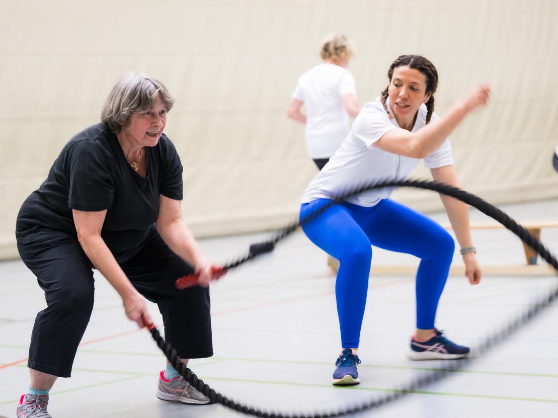 Zwei Frauen trainieren mit einem Battle Rope in einer Sporthalle, mehrere weitere Personen im Hintergrund aktiv.