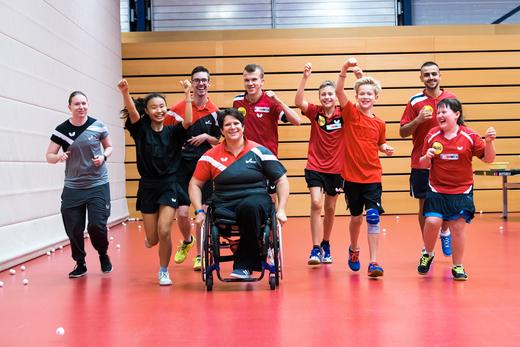 Gruppe von Tischtennisspielern in Sportkleidung läuft lächelnd und jubelnd auf rotem Boden in einer Sporthalle.