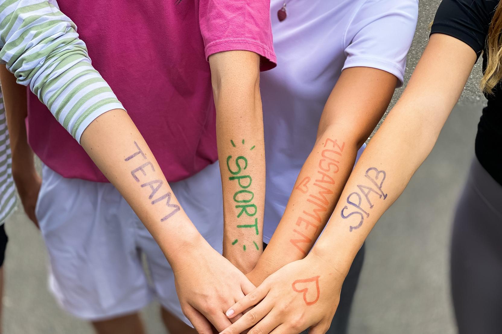 Vier Arme liegen übereinander, mit farbiger Schrift: „TEAM“, „SPORT“, „ZUSAMMEN“, „SPASS“ auf der Haut.