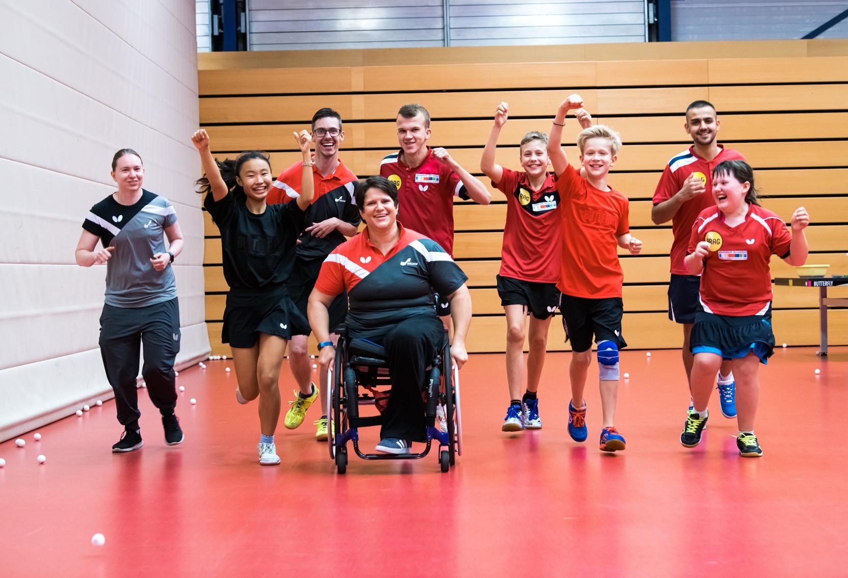 Gruppe von Tischtennisspielern in Sportkleidung läuft lächelnd und jubelnd auf rotem Boden in einer Sporthalle.