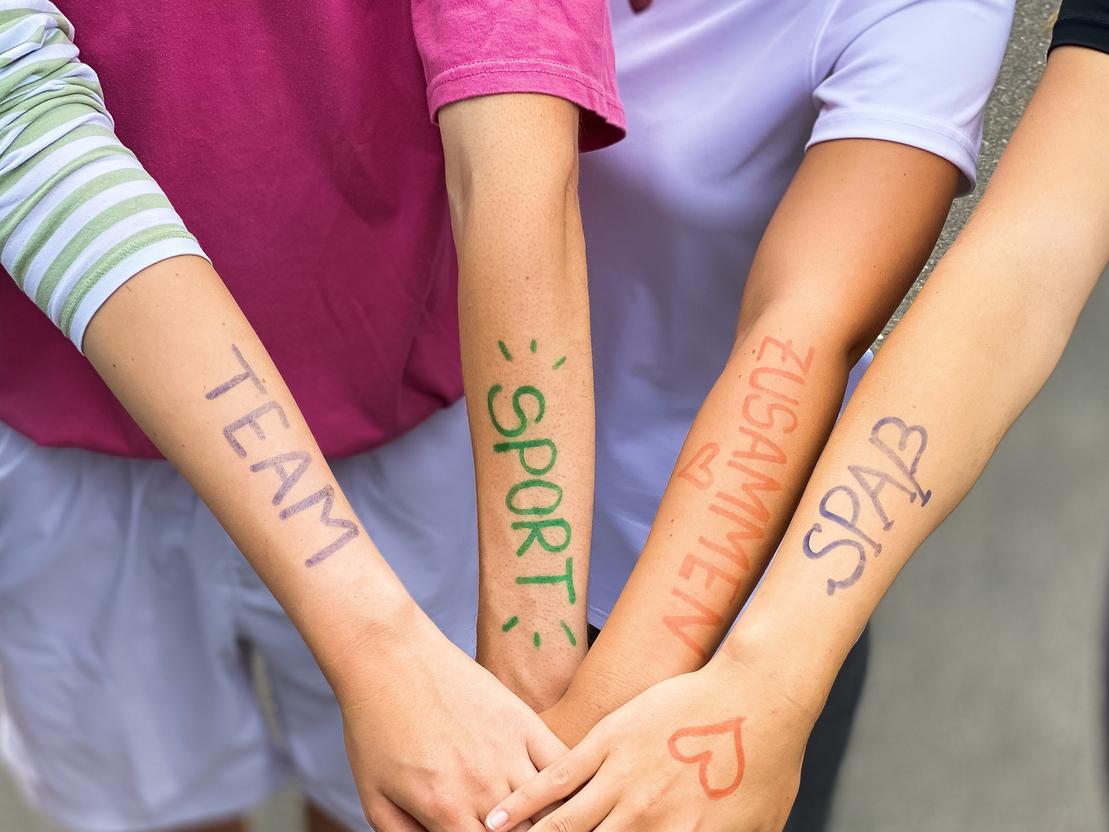 Vier Arme liegen übereinander, mit farbiger Schrift: „TEAM“, „SPORT“, „ZUSAMMEN“, „SPASS“ auf der Haut.