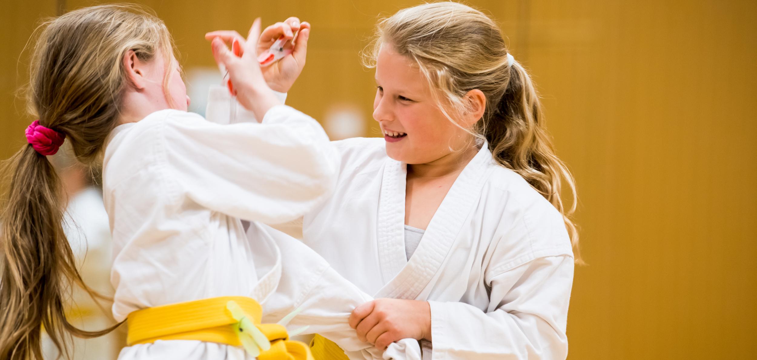 Zwei Mädchen in weißen Karateanzügen mit gelben Gurten üben freundschaftlich im Trainingsraum miteinander.