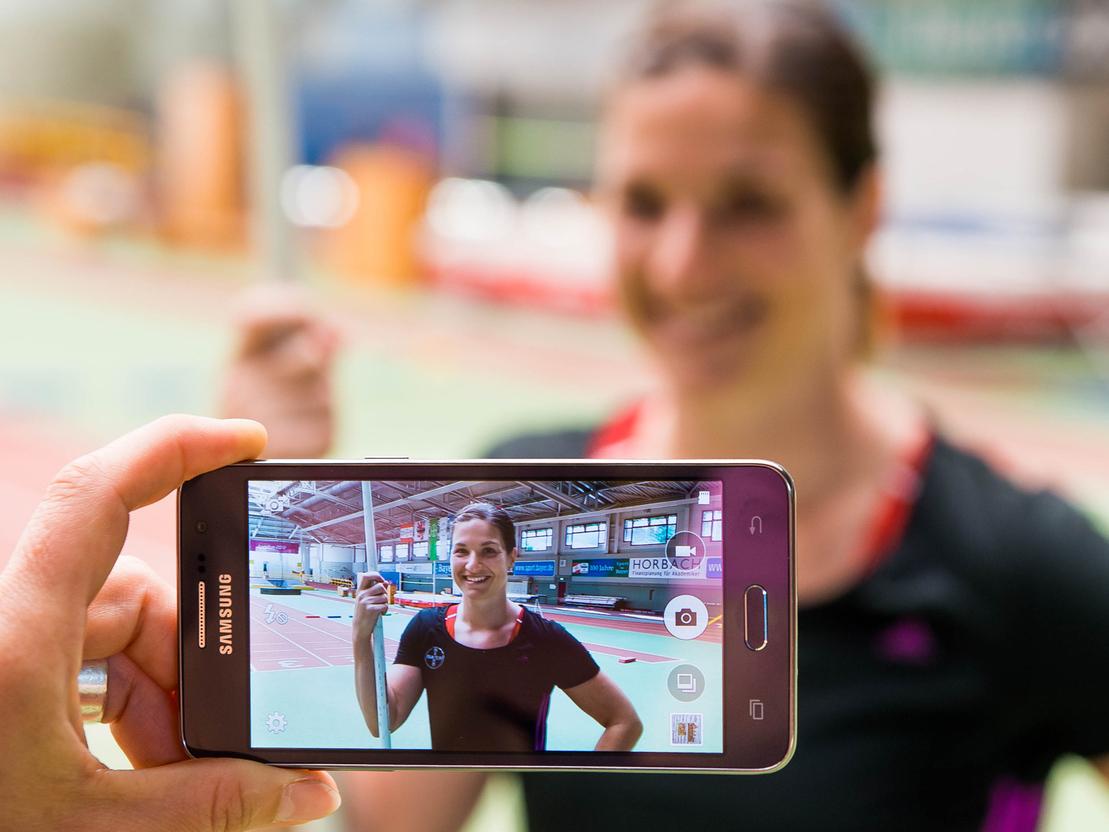 Hand hält ein Smartphone, das ein Porträt einer lächelnden Athletin mit Stab in einer Sporthalle zeigt.