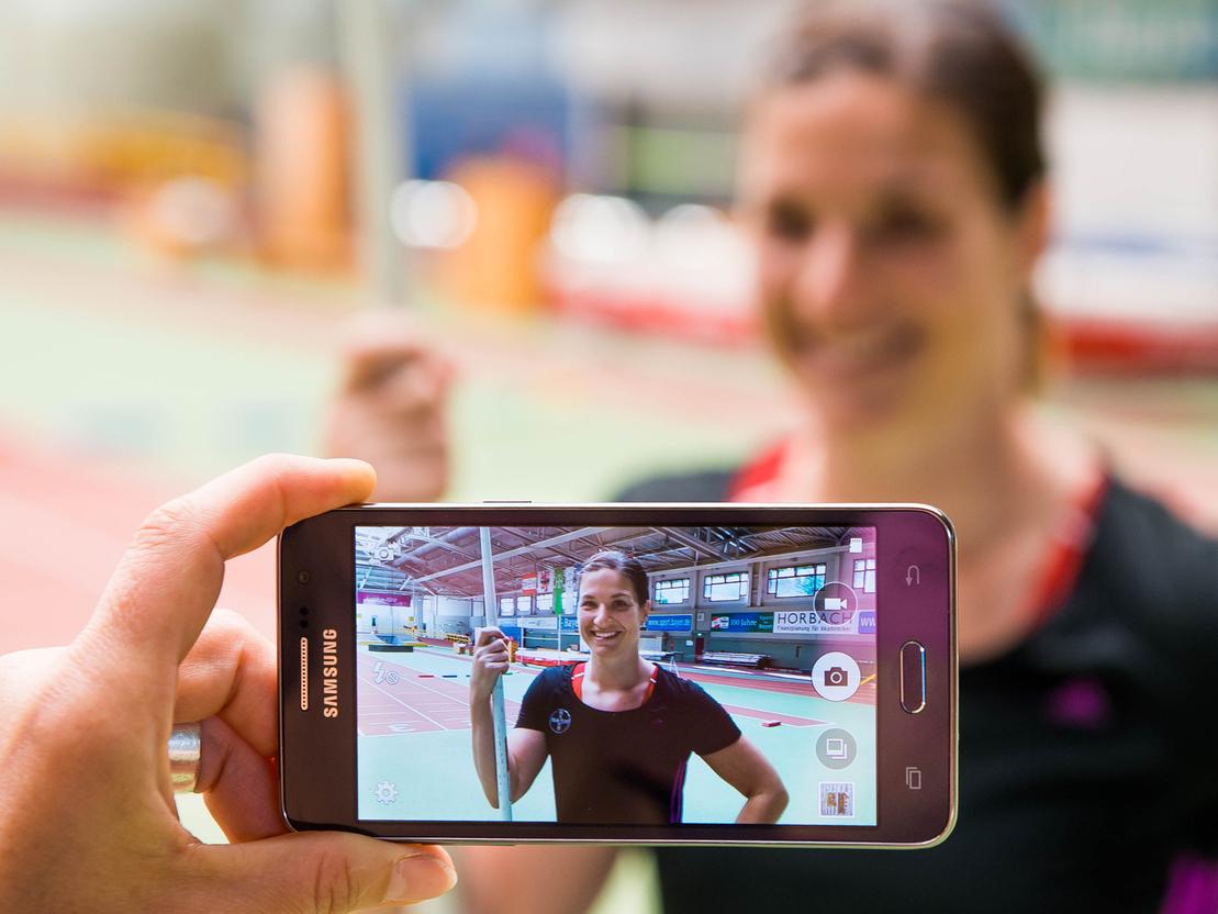 Hand hält ein Smartphone, das ein Porträt einer lächelnden Athletin mit Stab in einer Sporthalle zeigt.
