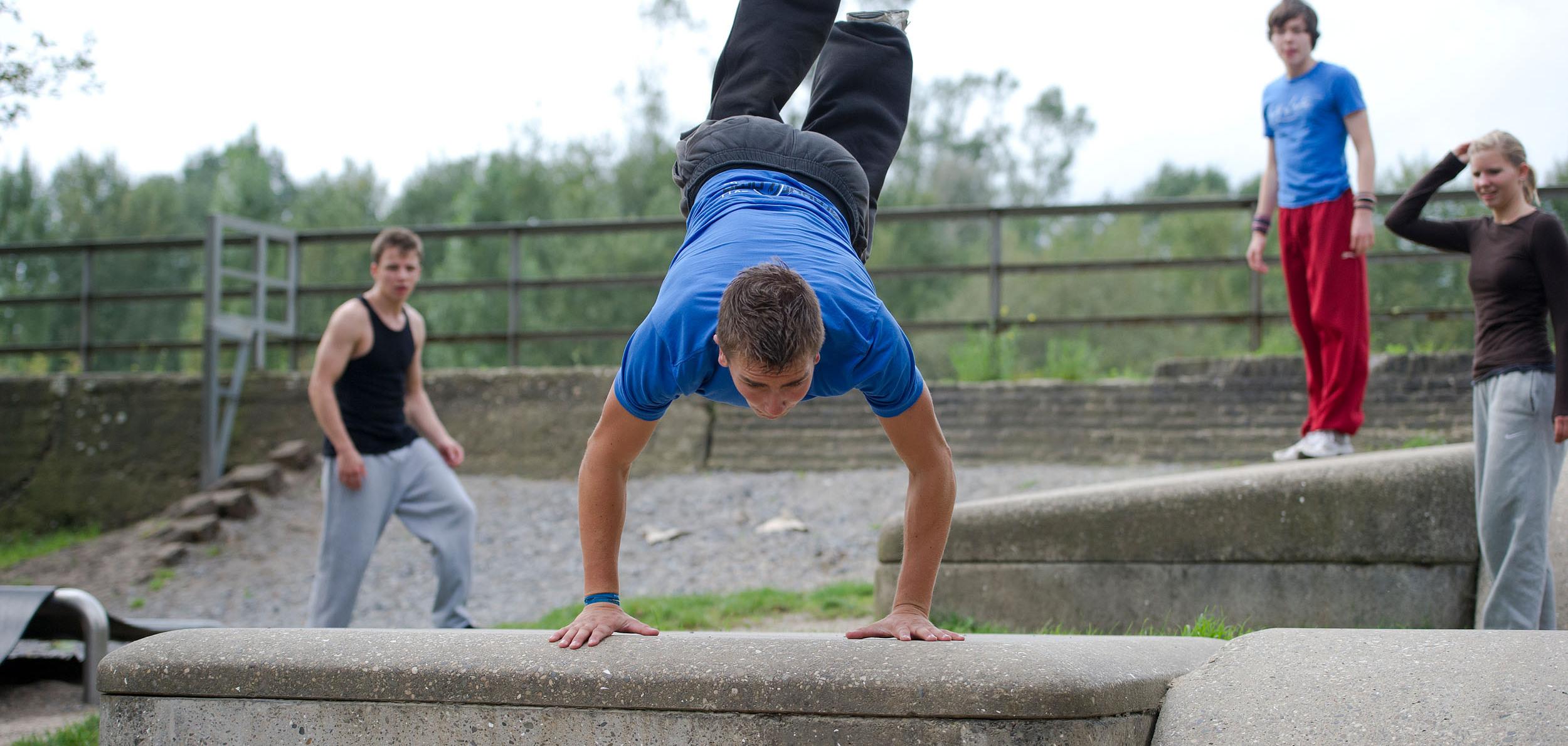Junger Mann im blauen T-Shirt führt einen Handstand auf einer Betonmauer aus, während andere zuschauen.