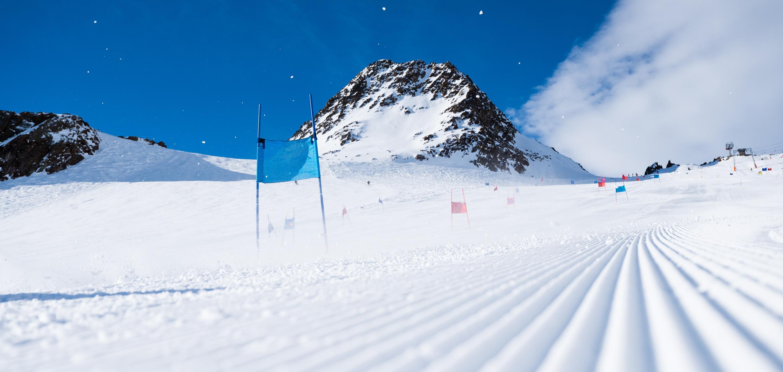 Schneebedeckte Piste mit blauen und roten Toren, im Hintergrund ein schneebedeckter Berg unter blauem Himmel.