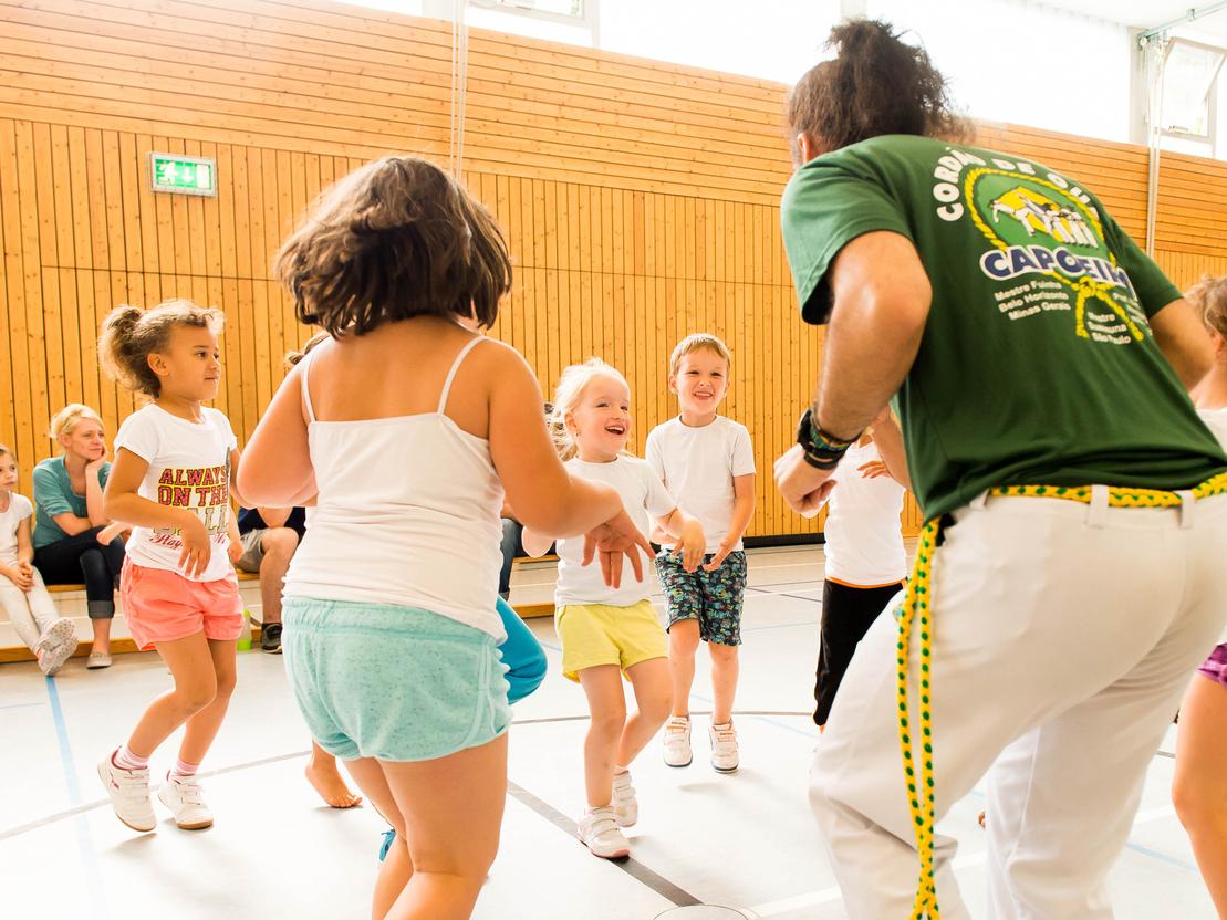 Gruppe von Kindern tanzt fröhlich in einer Turnhalle, geleitet von einem Trainer in grünem T-Shirt.