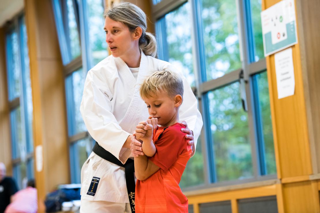 Eine Trainerin in Karate-Uniform zeigt einem Jungen mit rotem Shirt eine Technik im Dojo.