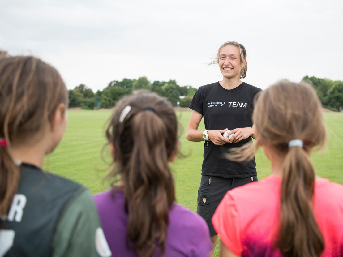 Trainerin spricht mit jungen Sportlerinnen auf einem grünen Fußballfeld unter bewölkten Himmel.