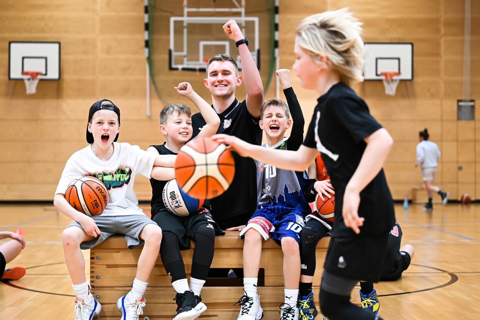 Gruppe fröhlicher Kinder und Trainer in Basketballkleidung posieren auf einer Bank in einer Sporthalle, mit Basketballs in den Händen.
