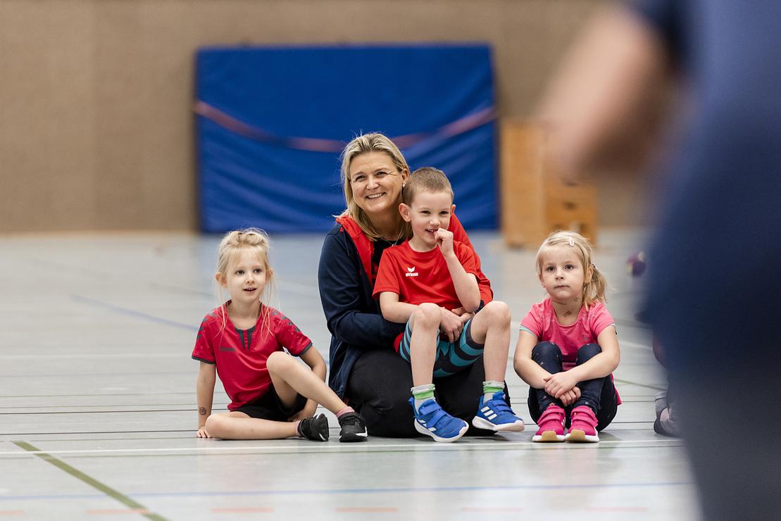 Eine Trainerin sitzt mit drei Kindern in Sportkleidung auf dem Boden in einer Sporthalle.