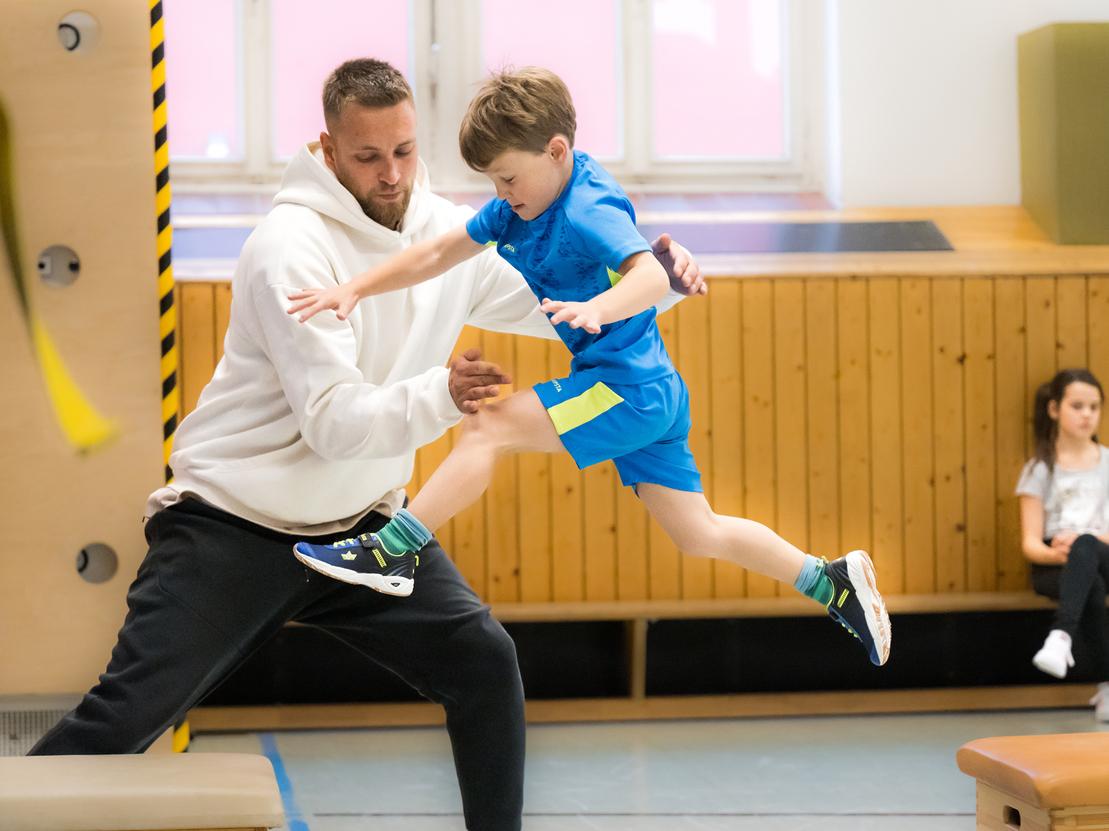 Ein junger Junge springt mit Hilfe eines Trainers über eine Trainingsbank in einer Turnhalle. Ein Mädchen beobachtet.