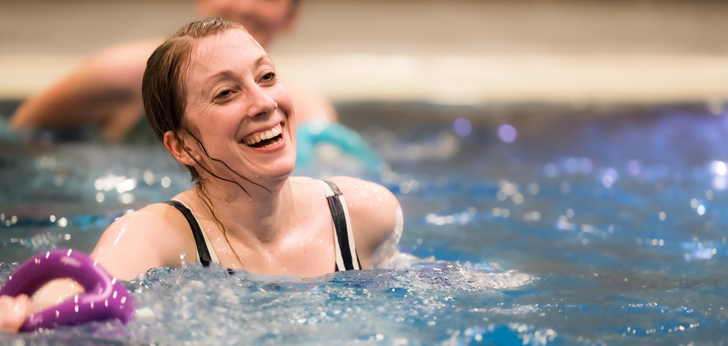 Lächelnde Frau im Wasser hält einen lila Schwimmring, im Hintergrund sind weitere Schwimmer zu sehen.